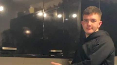 Police Scotland Cole Cooper, who is wearing a dark hoodie, looks at the camera while standing beside black kitchen cupboards. There are lights reflected in the cupboards.