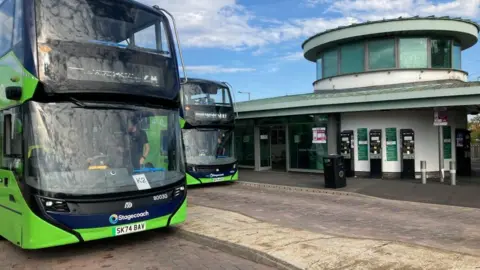 Two green buses are picking up passengers at Oxford Parkway Park and Ride. There is a circular building behind them with three ticket machines.