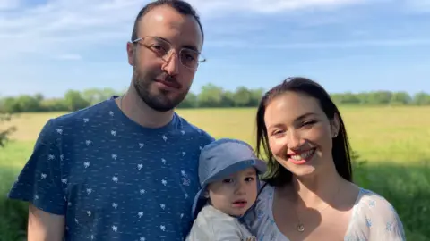 Kianty and Conor smile at the camera. They hold Luke, who wears a blue sunhat. There is a yellow field and a hedgerow in the background. 