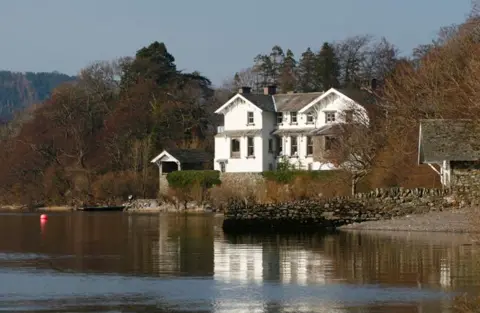 A view of Sharrow Bay taken from the lake. It is a white two-storey building with a slate roof on the shores of Ullswater. It is surrounded by trees.