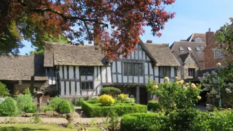 The Almonry Evesham The front of a large black and white timbered building with a green garden in front of it and a tree with red leaves overhanging it