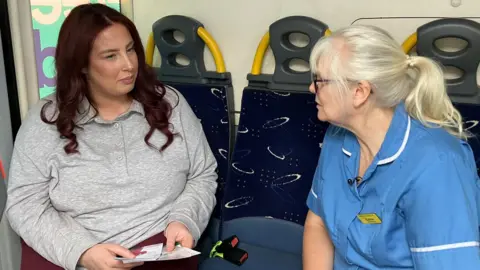 Two women sit on a bus in conversation. The woman on the left wears a grey long-sleeved polo top and holds some leaflets. She has long, curly, red-tinted hair. The woman on the right wears a nurses uniform and has white blonde hair tied back in a ponytail, a fringe, and glasses.