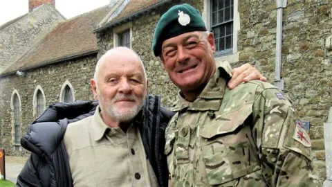 Sir Anthony Hopkins, left, standing next to Nick, right. Nick has a military uniform and beret on and Anthony Hopkins has a black coat on.