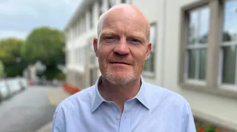 BBC Deputy Gavin St Pier - A bald man with a ginger a grey beard, wearing a buttoned up blue shirt with the collar open. 