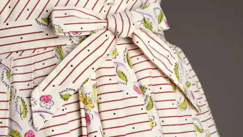 The Harris Close-up of the waist of a cotton dress with a belt tied in a bow. It is cream cotton with a fine red stripe 1cm apart with tiny black dots spaced widely in the lines and small green leaf and pink and yellow flowers dotted about.