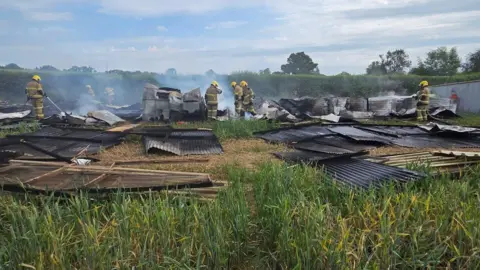 Shropshire Fire and Rescue Service Seven firefighters in beige operational kit and yellow helmets spray water onto charred and twisted corrugated metal in a field. Smoke is still rising from the buildings' remains.
