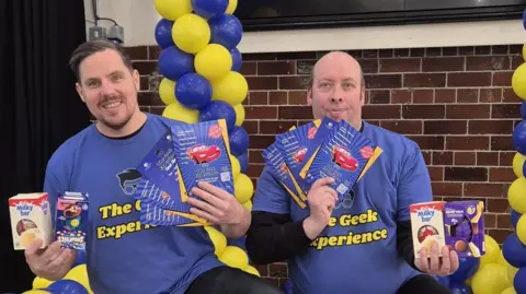 Luckbox Mike Brown and Mark Allison kneeling, holding leaflets and Easter eggs. They are both wearing purple t-shirts with yellow writing reading "The Geek Experience". There are yellow and blue balloons behind them. 