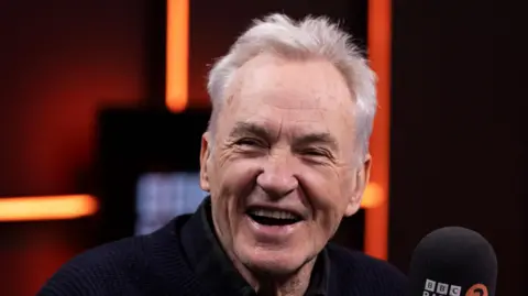 Larry Lamb, a man smiling at the camera. He is sitting next to a microphone.
He is wearing a black top. 