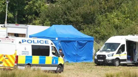 Eddie Mitchell A blue tent stands beside police and support vehicles near the Lewes derailment site. Emergency and investigation teams are working in a grassy area next to trees and railway infrastructure.