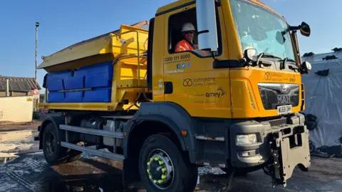 Staffordshire County Council A yellow gritting lorry with Staffordshire County Council written on the side. A male driver in an orange high-vis jacket and white hard hat is sitting in the front. The sky is blue in the background and the lorry is parked in a yard.