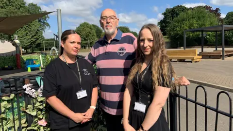 Three people are stood by railings at a children's nursery with a school playground in the background. A woman on the left has black hair and a black t-shirt, in the middle is a man with a grey beard and glasses wearing a pink and navy striped t-shirt and on the right is a woman with long wavy dark blond hair in a black t-shirt