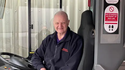 Paul Critchley, director of Blackburn Private Hire, has short grey hair and is wearing a navy blue fleece. He smiles as he sits behind the wheel of one of his company's electric buses.