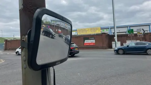 A convex mirror strapped to a lamppost on a traffic island at the junction of Lady Bay Bridge and Meadow Lane. Cars can be seen in the reflection of the mirror.