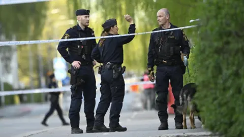 Getty Images Two policemen and a policewoman stand in black uniform with a dog between security tape on a pavement