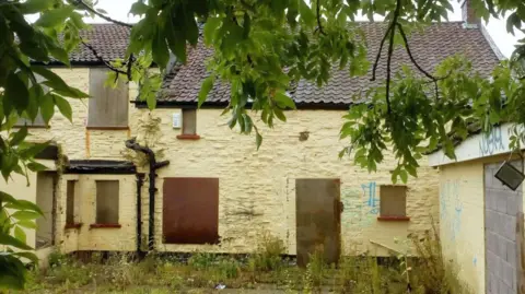Google The derelict pub photographed from inside the courtyard. It shows a yellowing building with boarded up windows and doors, and an overgrown patio.