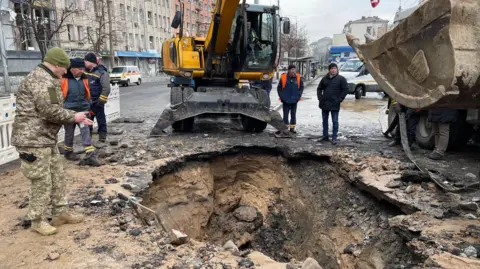 Emergency responders and a member of the military inspect a crater in the middle of a road, while another man operates a digger.