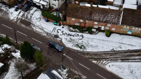 Ant Saddington/BBC An aerial image of village. On the ground in front of a row house is snow with the words "it's snowing" written it. A car is a travelling on the road and footprints can also been seen in the snow