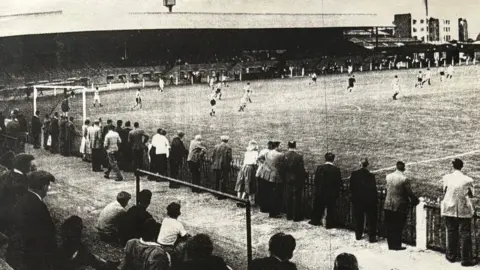 Tooting & Mitcham United FC A black and white photo of a football ground, seen from one corner looking across to approximately halfway along the pitch. Along the far side runs a covered terrace, with players on the pitch running away from the photographer's angle. In the foreground of the image is some uncovered terracing, with people sitting and standing on it.