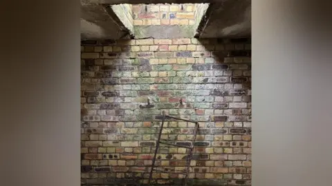 Keele University A brick wall inside a bunker with an escape hatch, leading to daylight. Below the hatch is an old metal ladder which is resting against the wall.