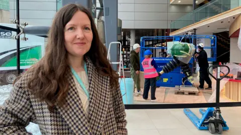 A woman with long, brown hair wearing a hound-tooth checked coat standing in front of as dark blue scissor lift and light-blue pallet truck with three people all wearing white or blue hard hats   