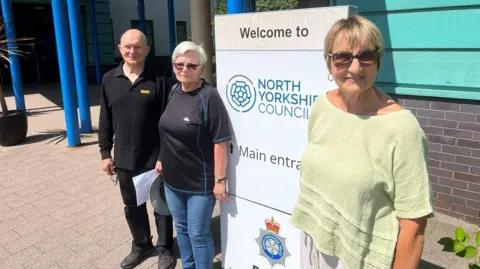 BBC/Richard Edwards Three people - two women and a man - stand outside a North Yorkshire Council building. The woman closest to the camera is wearing sunglasses and a light green top. The woman to the left of the picture is wearing sunglasses and a black top. The man is bald and is wearing a black top.