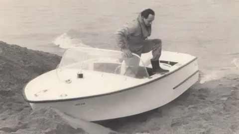 A black and white photograph of a speedboat docked on a sandbank on the edge of a large lake. Donald Campbell is stepping out of the boat on to the bank. 