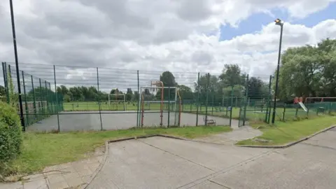 A play area surrounded by high green fences with football goals and basketball hoops at opposite ends. There is a paved area in the foreground and a playing field behind surrounded by trees. To the right is a playground area with a slide and other equipment.