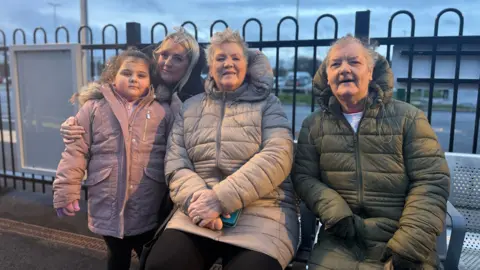 Christine Eastlake, in white jacket in the middle of the photo. She is sitting on a bench next to a women in a green jacket. A child in a pink jacket stands next to the bench as a well as a woman with blonde hair.