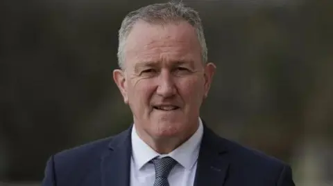 PA Conor Murphy in a black suit with a white shirt and polka dot tie.