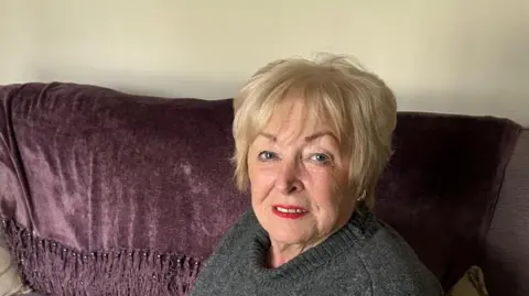 Shows a woman in blond hair with red lipstick and a grey round-neck jumper sitting on a sofa with a purple throw on top.