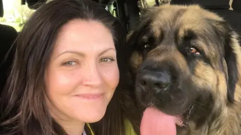A close-up of a woman with brown hair, and a dog with a large face sticking its tongue out. They appear to be in a car or minivan.