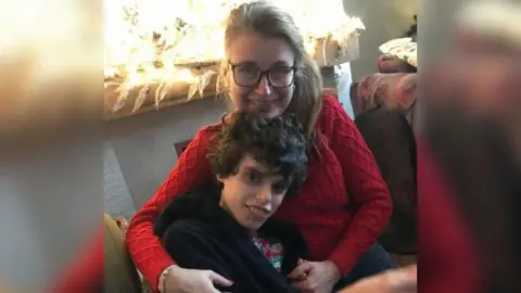 Family handout A woman with dark blonde hair and glasses wearing a red jumper hugs her son. He is wearing a Christmas top and a navy blue dressing gown. They are both smiling at the camera.