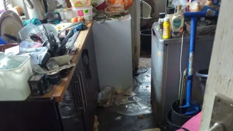 RSPCA The image depicts a narrow kitchen space with countertops on both sides and a doorway leading to another room in the background.
The floor is heavily littered with trash, food packaging, and miscellaneous items.