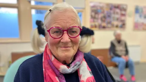 An elderly women is smiling at the camera. She has pink framed round glasses on and a pink and purple scarf. She has her hair in pigtails with a black scrunchies. 