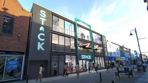 Artist's impression of the Stack venue at Bishop Auckland. It is a dark grey building with large glass windows. The Stack logo runs down both sides of the building as well as being above the entrance. Shoppers are walking along the street in front of it.