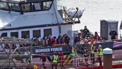 PA Media Channel migrants step onto the dock from a UK Border Force boat in Dover, Kent. The picture shows several migrants, wearing life jackets, being escorted from a boat by several officials wearing high-vis jackets.