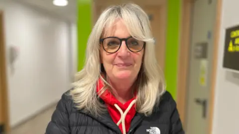 Karen Wagstaff, in a red hoodie under a black North Face coat, wearing glasses with medium length blonde hair - she is standing in a hospital corridor with a lime green door behind her.