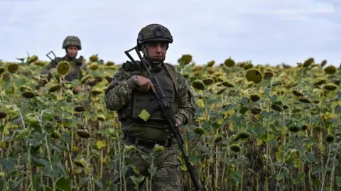 Reuters Two Ukrainian soldiers stand in a field of sunflowers near Pokrovsk - both are dressed in military gear and helmets, with the man in the foreground holding what looks to be a large machine gun 