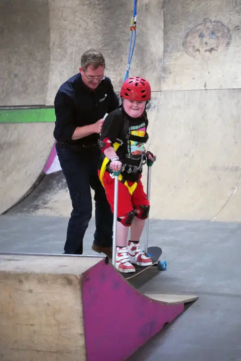 Cowshed A man holds onto a harness attached to a boy on a skateboard. He is wearing a red helmet and a black top with a colourful top on. A skateboard ramp can be seen in the background