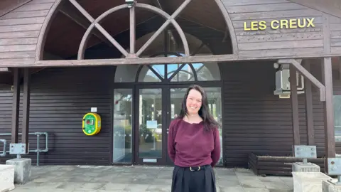 Jennifer Heald pictured outside Les Creux pavilion. She is wearing a plum coloured top and and is smiling. She has dark brown hair. In the background, the entrance to Les Creux is visible. The pavilion is mostly made of wood. 