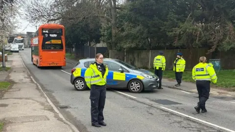 Four police officers in hi-viz jackets stand in the road near a police car. Further down the road is a large orange bus, the upper deck of which has been left misshapen due to damage. 