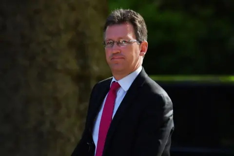 Getty A man walks outside on a sunny day. He has dark brown hair and glasses. He is wearing a black suit jacket, a pale blue shirt, and a maroon tie. 