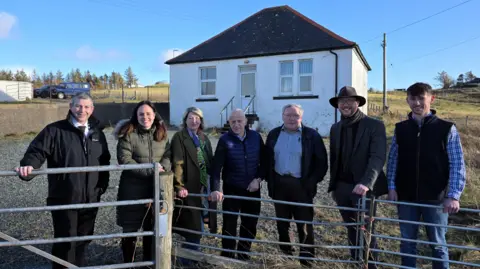 The former nurses' cottage is in the background. It is a small single storey white washed building with steps leading up to the front door. A group of people involved in the project are standing in the foreground next to a fence. 