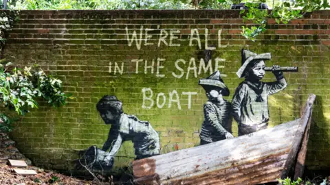 Banksy/PA Media A Banksy artwork, sprayed on the side of a small bridge, with three children wearing paper hats. One is looking through a telescope. Corrugated iron, made to look like a boat, has been placed next to the work to make it look at though the children were standing in it.