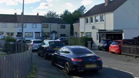 Google A cul-de-sac with brown and white houses. There are trees behind the houses at the far end. There are three cars in the foreground.