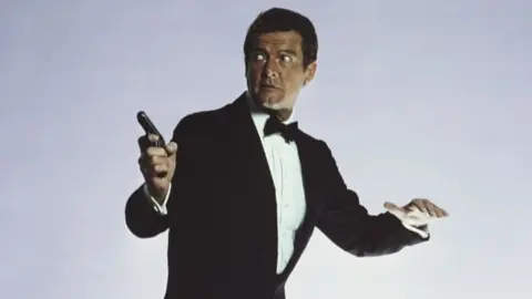 Roger Moore stars as James Bond. He is wearing a black dinner suit with white shirt and black bow tie. He is poised for action and carrying a pistol in his right hand. He is looking to his right.