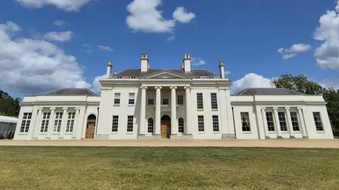 Stuart Woodward/BBC A large white building with columns has a grey roof and 4 chimney breasts on top of it. A lawn is in front and there is a blue sky above with some clouds and trees to the side.