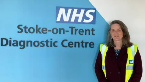 A woman with shoulder-length hair, wearing a high-vis vest over a dark jacket. She is stood alongside a large sign saying NHS Stoke-on-Trent diagnostic centre