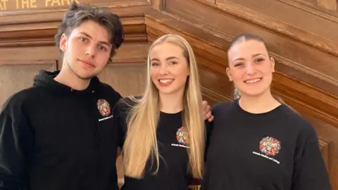 A teenage boy with medium-length brown hair and two teenage girls, one with long blonde hair and one with dark hair tied up in a ponytail. They're all wearing black tops branded with the Artemis College logo and are standing smiling, with their arms round each other, against a wood panelled wall.