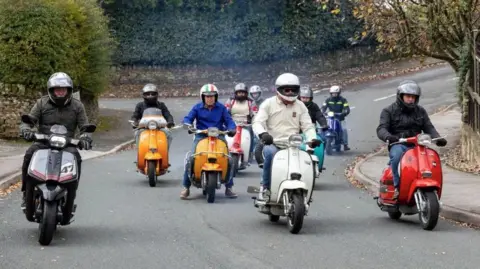 Bob Smith Several men on vintage mopeds riding down a street together. 